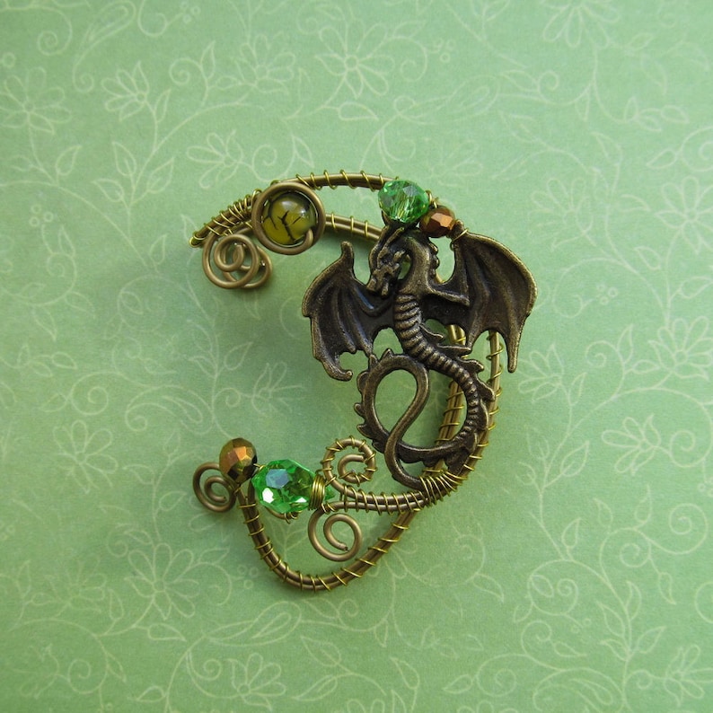 Puede incluir: Un pendiente de oreja de color bronce con un dise&ntilde;o detallado de drag&oacute;n. El pendiente est&aacute; adornado con cuentas verdes y amarillas, y un intrincado cableado. El drag&oacute;n tiene alas extendidas y una cola enroscada, un accesorio de fantas&iacute;a.