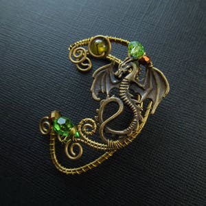 Puede incluir: Un pendiente de oreja de drag&oacute;n en tono bronce con detalles de gemas verdes y amarillas. El drag&oacute;n tiene alas detalladas y un cuerpo enrollado, la pieza est&aacute; hecha de alambre. Las gemas facetadas a&ntilde;aden color a la joya de fantas&iacute;a.