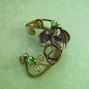 Puede incluir: Un pendiente de oreja de drag&oacute;n de color bronce con cuentas verdes y amarillas. El drag&oacute;n tiene alas detalladas y est&aacute; unido a un dise&ntilde;o envuelto en alambre. El pendiente de oreja est&aacute; sobre un fondo floral verde.