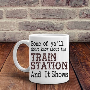 May include: White ceramic mug with a black handle. The mug features the text "Some of ya'll don't know about the TRAIN STATION And It Shows" in black and brown lettering. The mug is on a wooden surface.