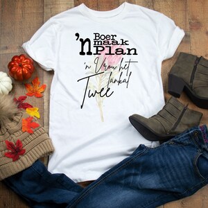 Peut inclure: T-shirt blanc avec texte en écriture noire et motif floral. Le t-shirt est associé à un jean bleu, des bottines vert olive et un bonnet en tricot marron avec un pompon en fausse fourrure. Des citrouilles décoratives et des feuilles d'automne sont également présentes.