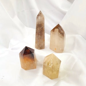 May include: Four clear quartz crystal points in various shades of brown and yellow. The crystals are arranged on a white surface.