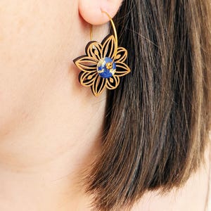 Puede incluir: Pendientes de aro dorados con un diseño de flor calada en láser marrón oscuro. La flor presenta un centro azul y dorado. Los pendientes se llevan en la oreja de una persona, mostrando los intrincados detalles y el contraste de color.