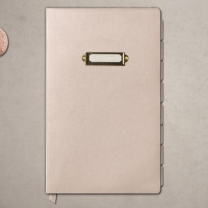 May include: A beige leather bound journal with a gold label on the front cover. The journal has a ring binding and a ribbon bookmark.