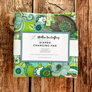 May include: A folded diaper changing pad featuring a vibrant green, teal, and brown floral paisley pattern. It is secured with a light green snap tab and wrapped in a white paper band labeled Willow Tree Crafting, 25x17 inches, and machine washable.