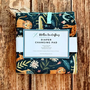May include: A folded dark green diaper changing pad with a colorful jungle animal print featuring lions and giraffes. A white paper band displays the text Willow Tree Crafting, Diaper Changing Pad, 25x17 inches, and other product details.