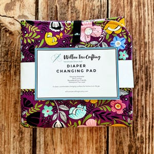 May include: A folded diaper changing pad with a purple background and a colorful floral pattern featuring yellow, pink, and teal flowers. A white paper label reads Willow Tree Crafting, DIAPER CHANGING PAD, Machine Washable, 25x17 inches, Reusable & Eco-Friendly, Waterproof Top Layer.