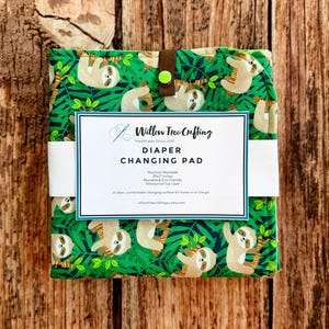 May include: A folded diaper changing pad featuring a green jungle pattern with sloths. A white paper band wraps around the center, printed with the text Willow Tree Crafting, Diaper Changing Pad, 25x17 inches, and other product details.