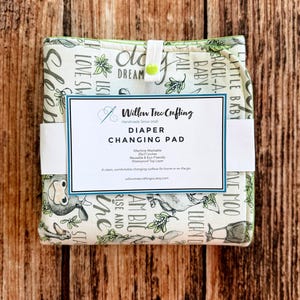 May include: A folded fabric diaper changing pad with a cream background featuring green illustrations of farm animals and text. A white paper label reads Willow Tree Crafting, Diaper Changing Pad, 25x17 inches, and other product details.