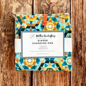 May include: A folded diaper changing pad with a geometric pattern in teal, yellow, orange, and dark blue. It features an orange snap closure and a white paper label reading Willow Tree Crafting, Diaper Changing Pad, 25x17 inches, and more.