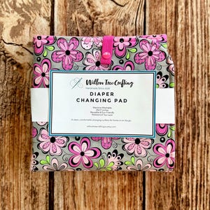 May include: A folded diaper changing pad with a gray background and a repeating pattern of pink, black, and light green flowers. A white paper band features the text Willow Tree Crafting, Diaper Changing Pad, 25x17 inches, and other details.