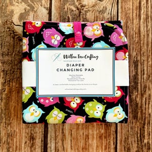 May include: A folded diaper changing pad featuring a black fabric base with a colorful pattern of pink, blue, and green owls. A white paper label is wrapped around the center with the text Willow Tree Crafting, Diaper Changing Pad, 25x17 inches.