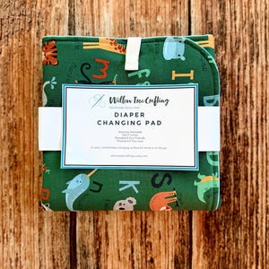 May include: A folded green diaper changing pad with a colorful animal and alphabet print. A white paper band features the text Willow Tree Crafting, Diaper Changing Pad, Machine Washable, 25x17 inches, Reusable & Eco-Friendly, Waterproof Top Layer.