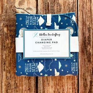 May include: A folded blue diaper changing pad featuring a celestial pattern of moons, stars, and clouds with the text 'SHOOT FOR THE MOON REACH FOR THE STARS'. A white paper band displays the text 'Willow Tree Crafting DIAPER CHANGING PAD'.