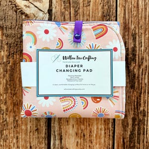 May include: A folded pink diaper changing pad featuring a pattern of colorful rainbows and white daisies. A purple snap closure secures the pad. A white label reads Willow Tree Crafting, Diaper Changing Pad, 25x17 inches, Machine Washable, Reusable & Eco-Friendly.