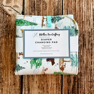 May include: A folded fabric diaper changing pad with a forest-themed print featuring green trees, brown bears, and foxes. A white paper label reads Willow Tree Crafting, Diaper Changing Pad, 25x17 inches, Machine Washable, Reusable & Eco-Friendly.