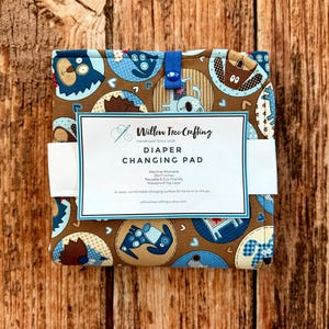 May include: A folded fabric diaper changing pad featuring a brown background with a repeating pattern of blue and brown cartoon dogs in circular frames. A blue elastic strap secures the pad, and a white product label reads Willow Tree Crafting.