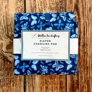 May include: A folded blue diaper changing pad with a white butterfly pattern and a snap closure. A white paper label reads Willow Tree Crafting, Diaper Changing Pad, Machine Washable, 25x17 inches, Reusable & Eco-Friendly, Waterproof Top Layer.