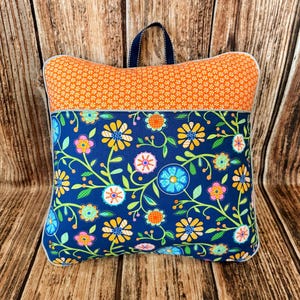 May include: A decorative pillow with a navy blue floral pattern and an orange top section. The pillow has a white trim and a navy blue handle. The floral design includes yellow, pink, and blue flowers with green leaves.