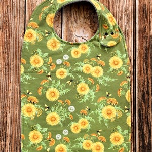 May include: A green baby bib with a pattern of yellow dandelions, white dandelion seeds, and black bees. The bib has a rounded neck and snaps for closure. The fabric is a soft, cotton material.