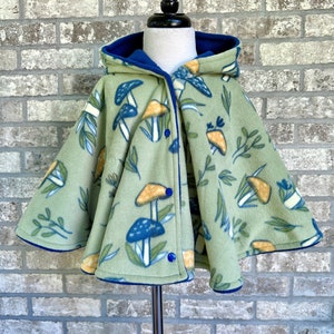 May include: A light green fleece cape with a mushroom and leaf pattern. The cape has a blue hood and trim, and fastens with blue buttons. The cape is displayed on a mannequin.
