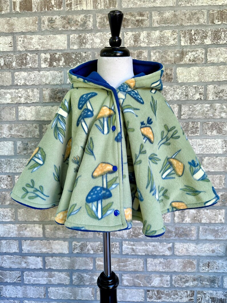 May include: A light green fleece cape with a mushroom and leaf pattern. The cape has a blue hood and trim, and fastens with blue buttons. The cape is displayed on a mannequin.