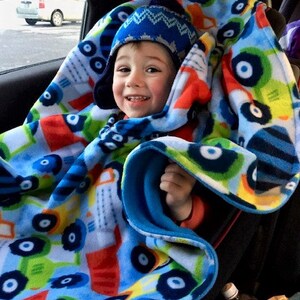 May include: A child wearing a blue knit hat is wrapped in a colorful fleece blanket. The blanket features a pattern of blue, green, red, and yellow vehicles. The child is seated in a car seat.