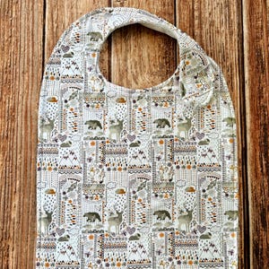 May include: A baby bib featuring a repeating pattern of deer, bears, mountains, hearts, and clouds in shades of brown, orange, and gray on a white background. The bib has a rounded neckline and a side closure with a fabric fastener.