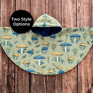 May include: A green fleece poncho with a hood, featuring a pattern of blue and yellow mushrooms and leaves. The hood has a dark blue lining. The text "Two Style Options" is in a black circle. The poncho is displayed on a wooden surface.
