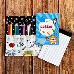 May include: A fabric pencil roll with a black star pattern and a white section featuring school-themed graphics, text, and colored pencils. Beside it is a blue notebook with a cartoon animal cover featuring the word Letter and alphabet letters.