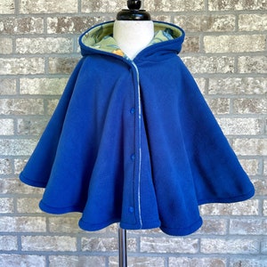 May include: A royal blue hooded cape with a floral patterned lining. The cape features a button closure down the front and is displayed on a mannequin. The cape is made of a soft material.