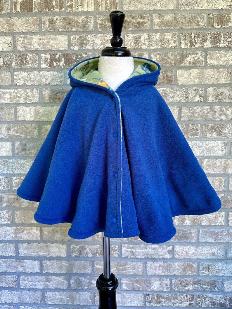 May include: A royal blue hooded cape with a floral patterned lining. The cape features a button closure down the front and is displayed on a mannequin. The cape is made of a soft material.