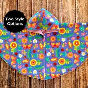 May include: A hooded cape featuring a purple background with a colorful pattern of orange, yellow, and pink flowers, red ladybugs, and butterflies. The garment has a light pink interior lining and a front closure with white snaps. Text reads Two Style Options.