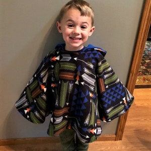 May include: A child wearing a black hooded poncho with a colorful arrow pattern in shades of green, orange, white, and blue. The poncho has a blue lining and is paired with green pants and white socks.