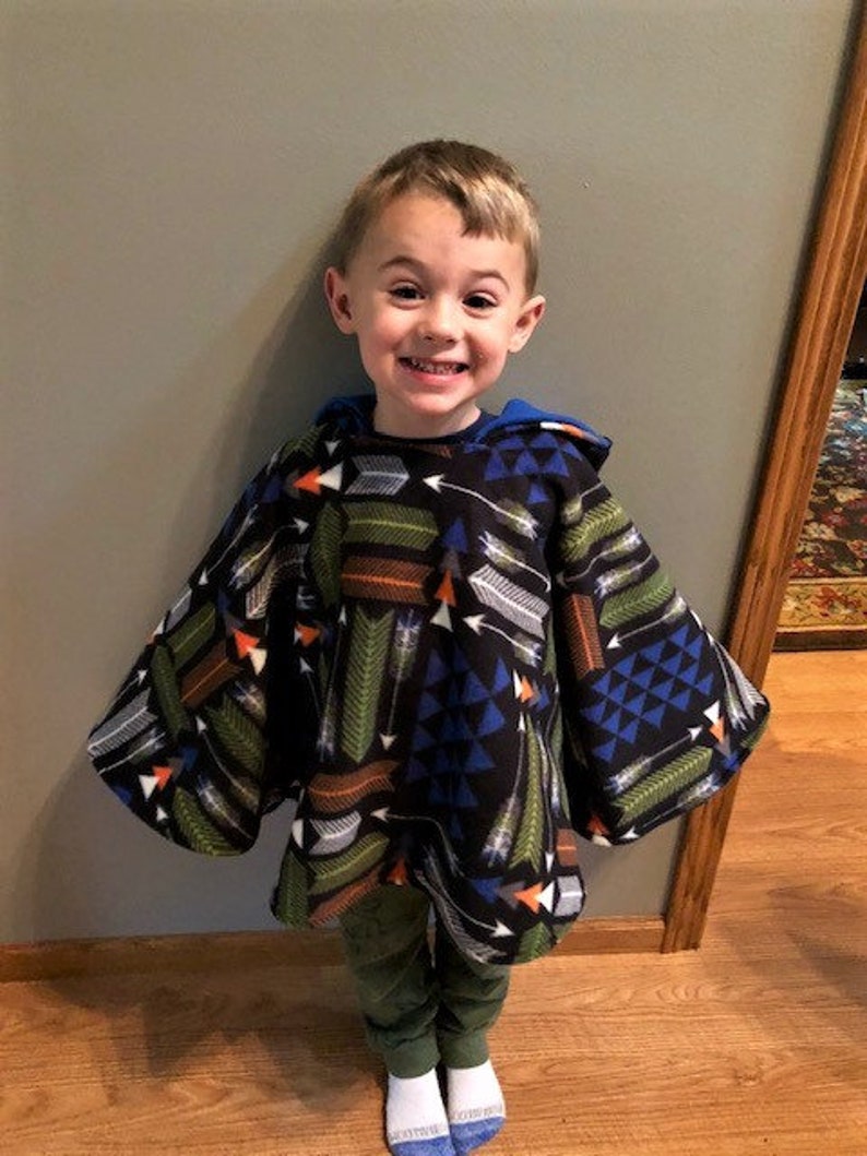 May include: A child wearing a black hooded poncho with a colorful arrow pattern in shades of green, orange, white, and blue. The poncho has a blue lining and is paired with green pants and white socks.