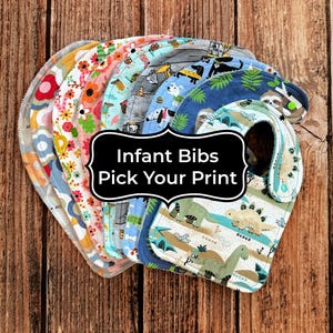May include: A stack of colorful infant bibs with various patterns, including floral, animal, and geometric designs. The bibs are made of soft fabric and have a curved shape to catch spills. Text on the image reads "Infant Bibs Pick Your Print."