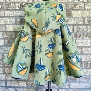 May include: A light green fleece hooded cape with a mushroom and leaf pattern. The mushrooms are blue and orange, and the leaves are green. The cape has a blue trim and is displayed on a black mannequin.