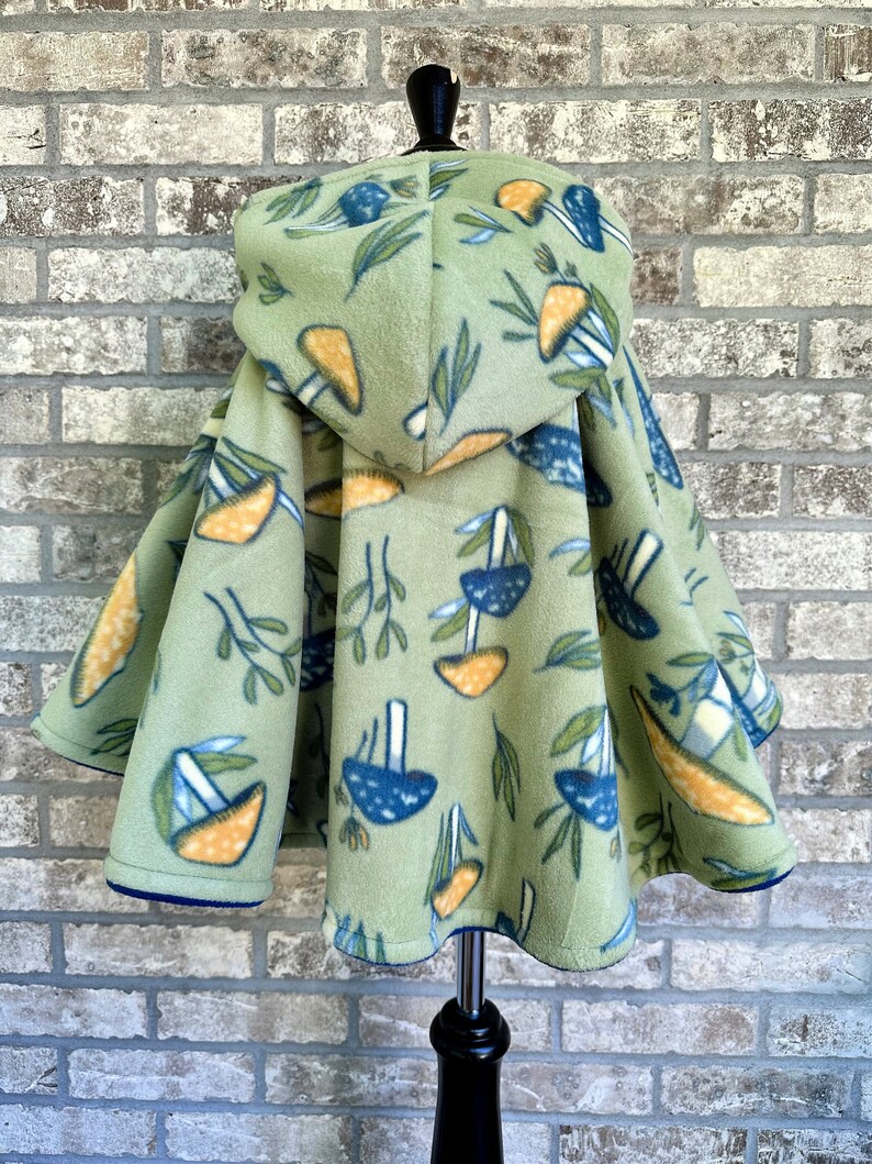 May include: A light green fleece hooded cape with a mushroom and leaf pattern. The mushrooms are blue and orange, and the leaves are green. The cape has a blue trim and is displayed on a black mannequin.