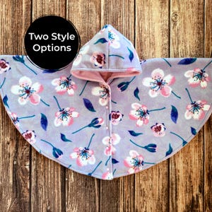 May include: A light purple fleece hooded cape featuring a repeating pattern of pink and white cherry blossoms with dark blue leaves. The garment has a hood, a front opening with small white buttons, and the text Two Style Options in a black circle.
