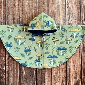 May include: A light green fleece poncho with a hood, featuring a repeating pattern of blue and yellow mushrooms and green leaves. The poncho has a dark blue lining and is buttoned down the front.