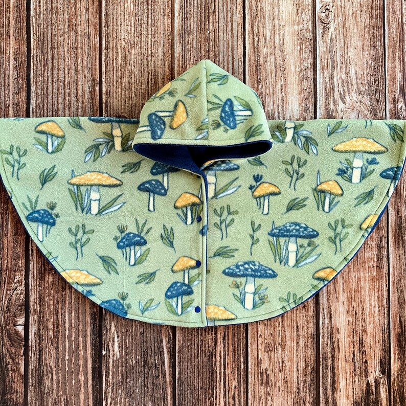 May include: A light green fleece poncho with a hood, featuring a repeating pattern of blue and yellow mushrooms and green leaves. The poncho has a dark blue lining and is buttoned down the front.