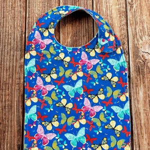 May include: A blue baby bib featuring a repeating pattern of butterflies in various colors including pink, yellow, turquoise, and red. The bib has a rounded neckline and a fabric fastener on the side, with a consistent print covering the entire surface.
