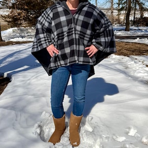 May include: A hooded cape in a black, gray, and white plaid pattern. The cape has a zipper closure and is worn over blue jeans and brown boots. The person is standing in the snow.