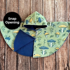 May include: A green fleece hooded cape with a mushroom pattern and a snap opening. The cape features a blue lining and trim. The hood and cape are decorated with illustrations of blue and yellow mushrooms and foliage. The text "Snap Opening" is in a black circle.