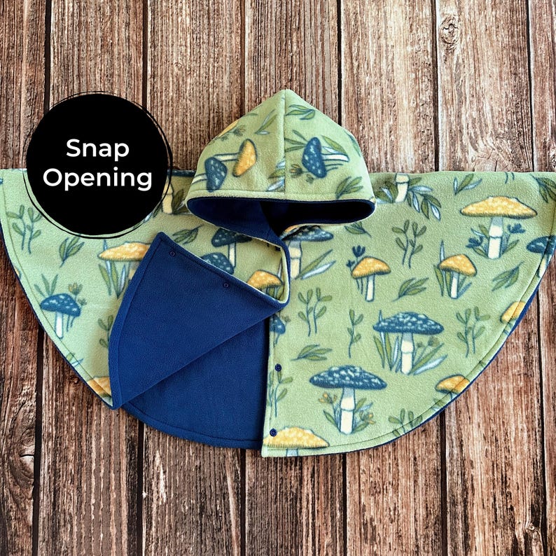 May include: A green fleece hooded cape with a mushroom pattern and a snap opening. The cape features a blue lining and trim. The hood and cape are decorated with illustrations of blue and yellow mushrooms and foliage. The text "Snap Opening" is in a black circle.