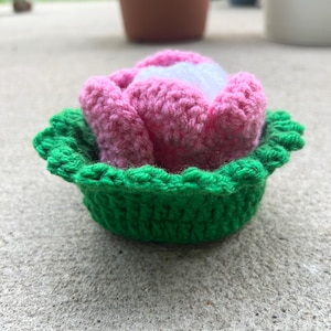 May include: A handmade crochet flower in a green bowl. The flower is pink with white in the center. The bowl is green and has a scalloped edge. The item is sitting on a concrete surface.