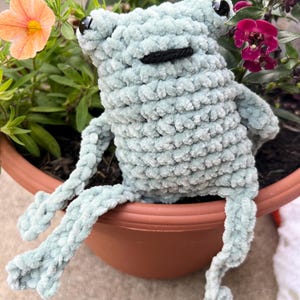 May include: A light blue crocheted frog with black eyes and a black mouth sits in a brown flower pot. The frog has long, dangling legs and is surrounded by green foliage and colorful flowers. The frog is made of a soft, textured yarn.
