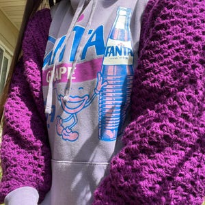 May include: A light purple hooded sweatshirt featuring a graphic of a Fanta Grape bottle and a cartoon character. The sleeves are made of a thick, dark purple crocheted material with a textured, open-knit pattern, attached to the sweatshirt body.