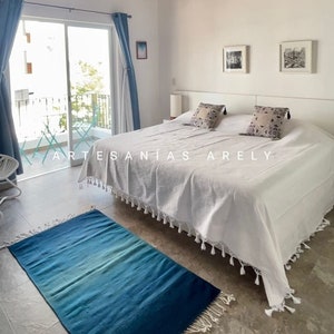 May include: A bedroom featuring a white bedspread, two patterned cushions, and a blue and teal rug. The room has a balcony with a view and blue curtains. The text "ARTESANIAS ARELY" is visible.