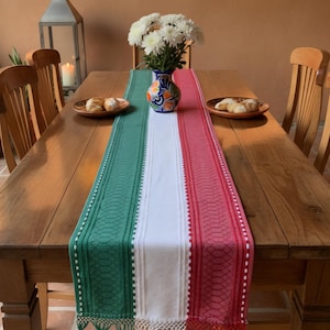 Peut inclure: Une table à manger en bois est ornée d'un chemin de table coloré. Le chemin de table présente des rayures verticales vertes, blanches et rouges, avec des glands décoratifs à chaque extrémité. Un vase de fleurs blanches est au centre, et des assiettes de nourriture sont de chaque côté.